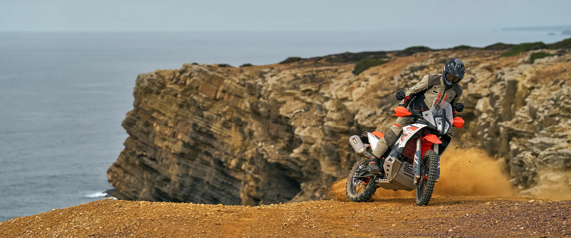 KTM 890 Adventure R en virage sur une piste côtière, falaise et océan en arrière-plan