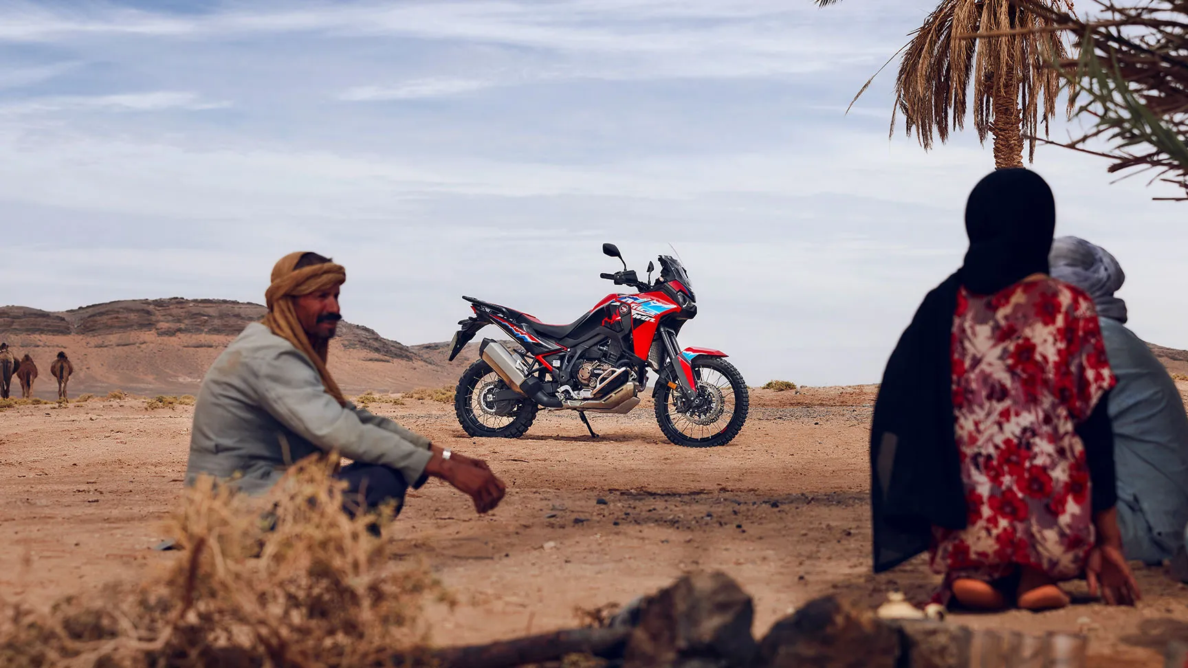 Honda CRF1100L Africa Twin à l'arrêt dans un paysage saharien, deux personnes assises au premier plan
